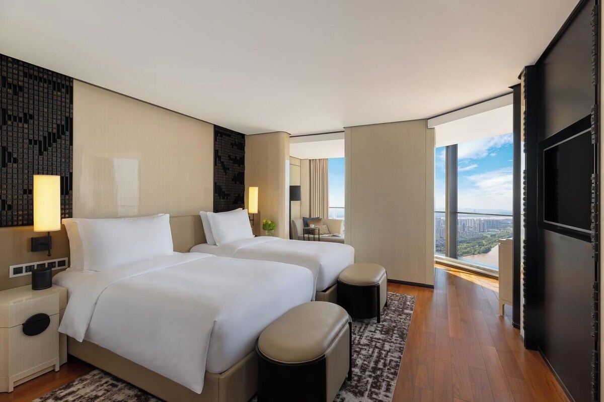 Une chambre d’hôtel moderne avec deux lits, une grande fenêtre donnant sur la ville, un téléviseur à écran plat et un petit coin salon.