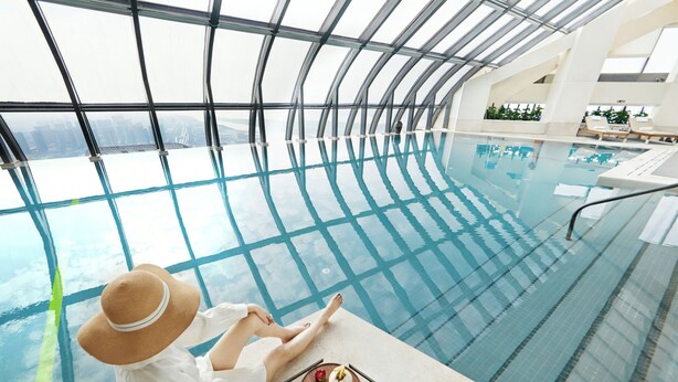 Eine Person sitzt am Pool mit Blick auf einen Stadtpanoramablick durch eine Glasdecke.