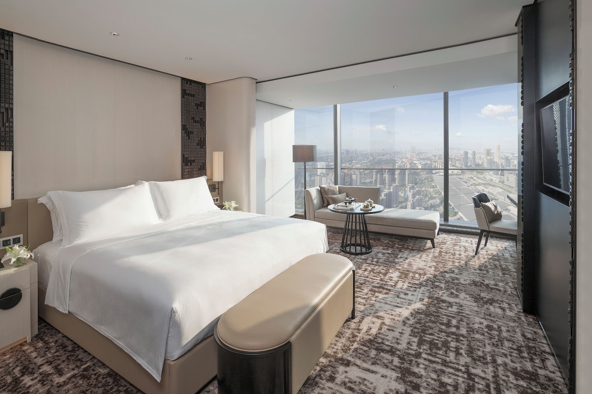 A modern hotel room with a large bed, a sofa, a small table, and a view of the city through large windows.