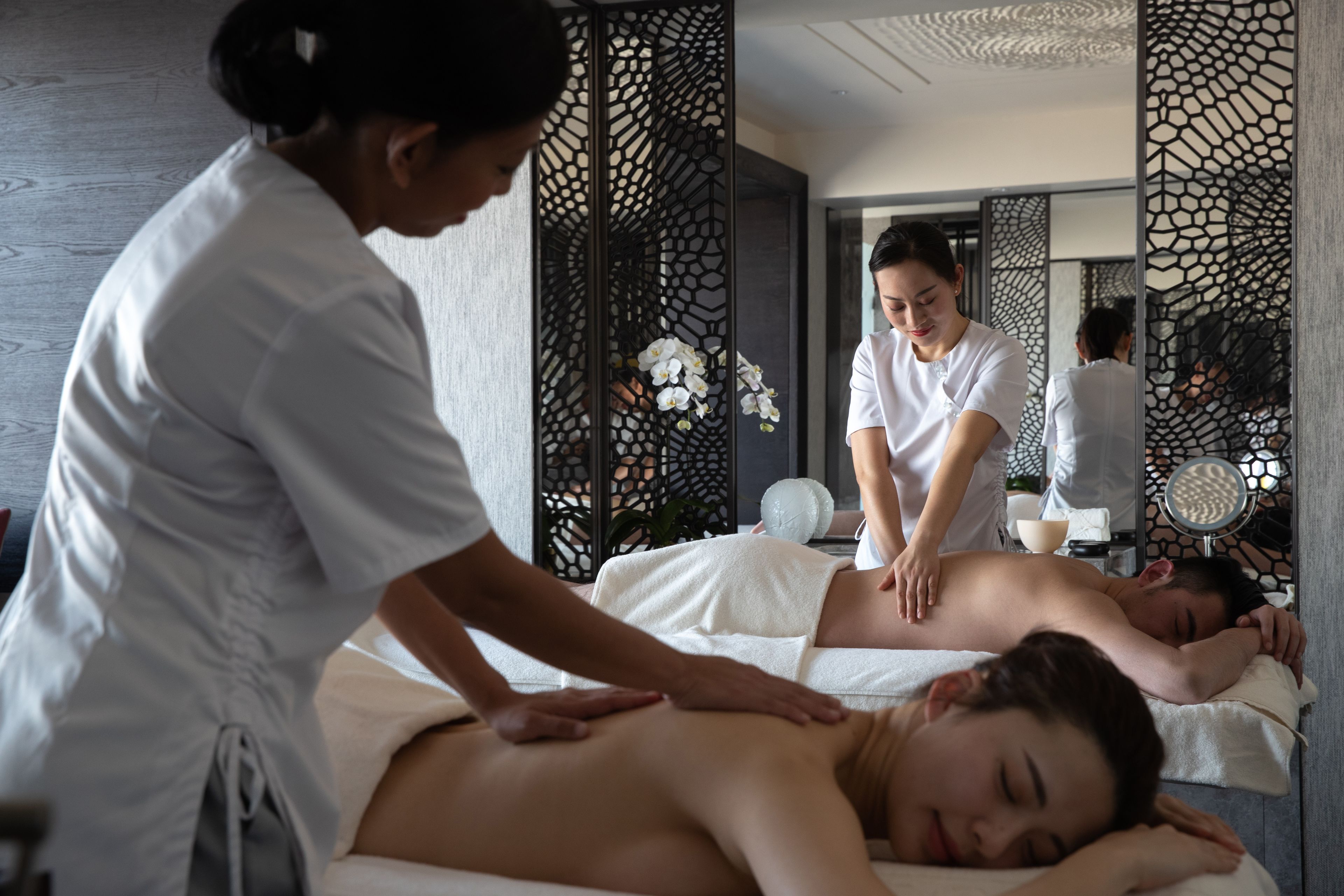 Un cuarto de spa con una camilla de masaje, una persona recibiendo un masaje en la espalda y otra de pie.