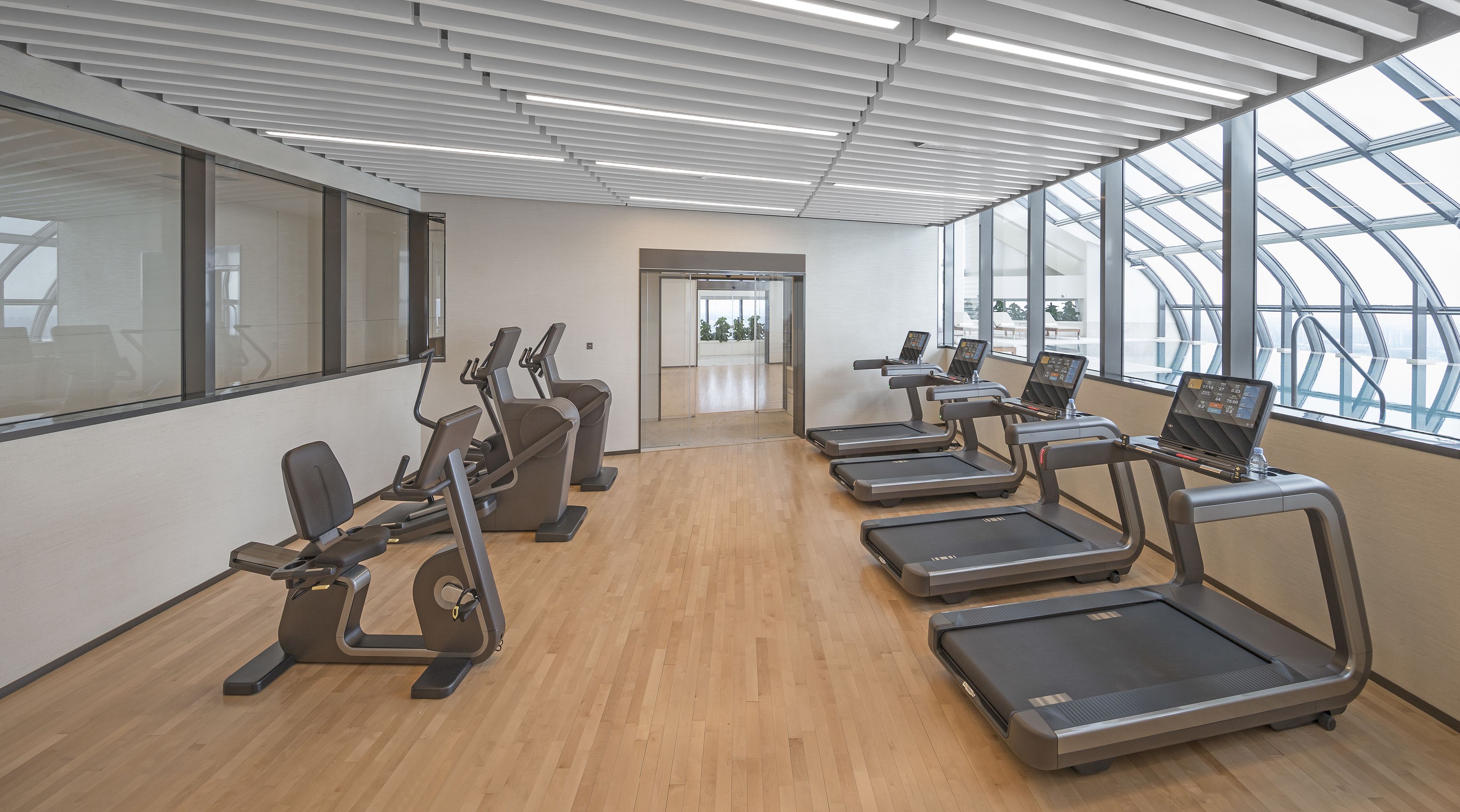 A modern gym with treadmills, elliptical machines, and a large glass window offering a view of the outdoors.