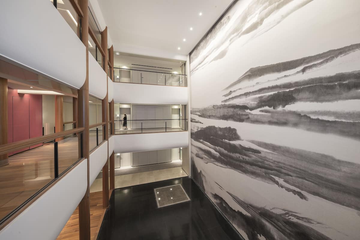 Un interior de varios niveles con un cuadro abstracto grande en la pared derecha, una escalera que une los pisos, y una persona caminando en el nivel superior.