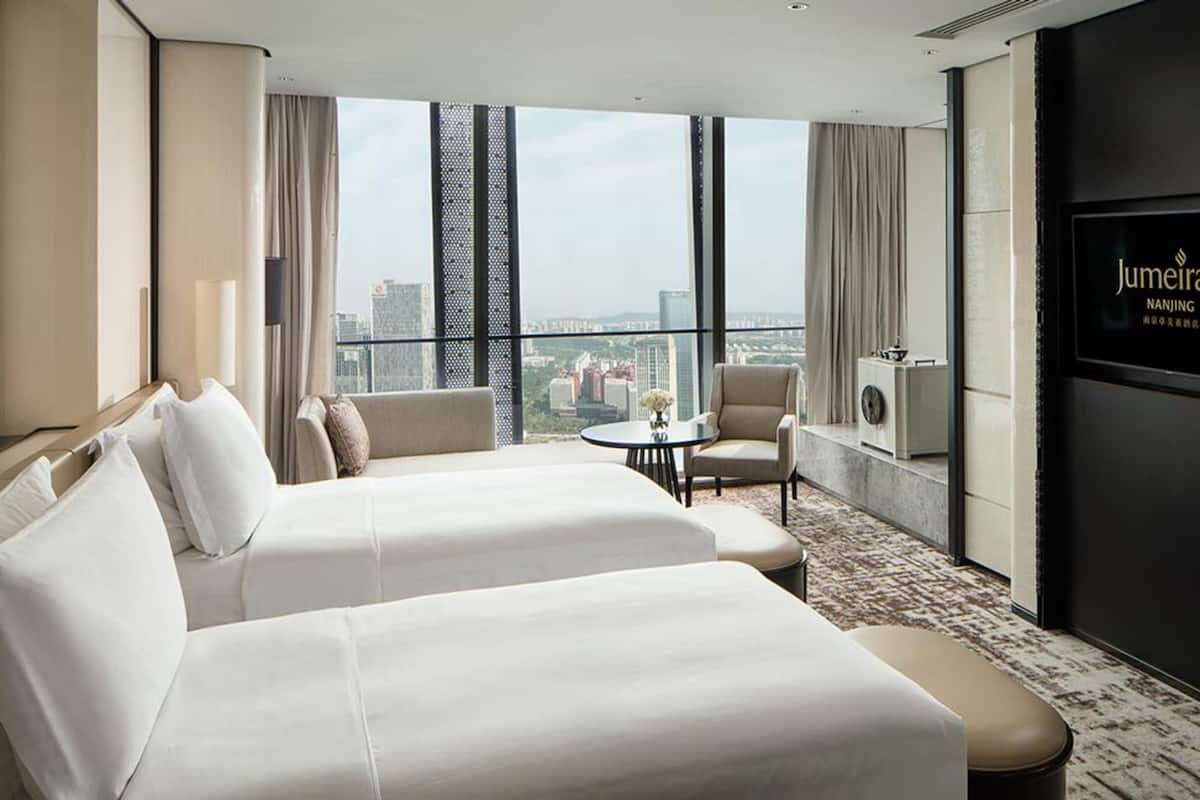 Habitación de hotel con dos camas, una mesita, una silla, televisor y vista a la ciudad.