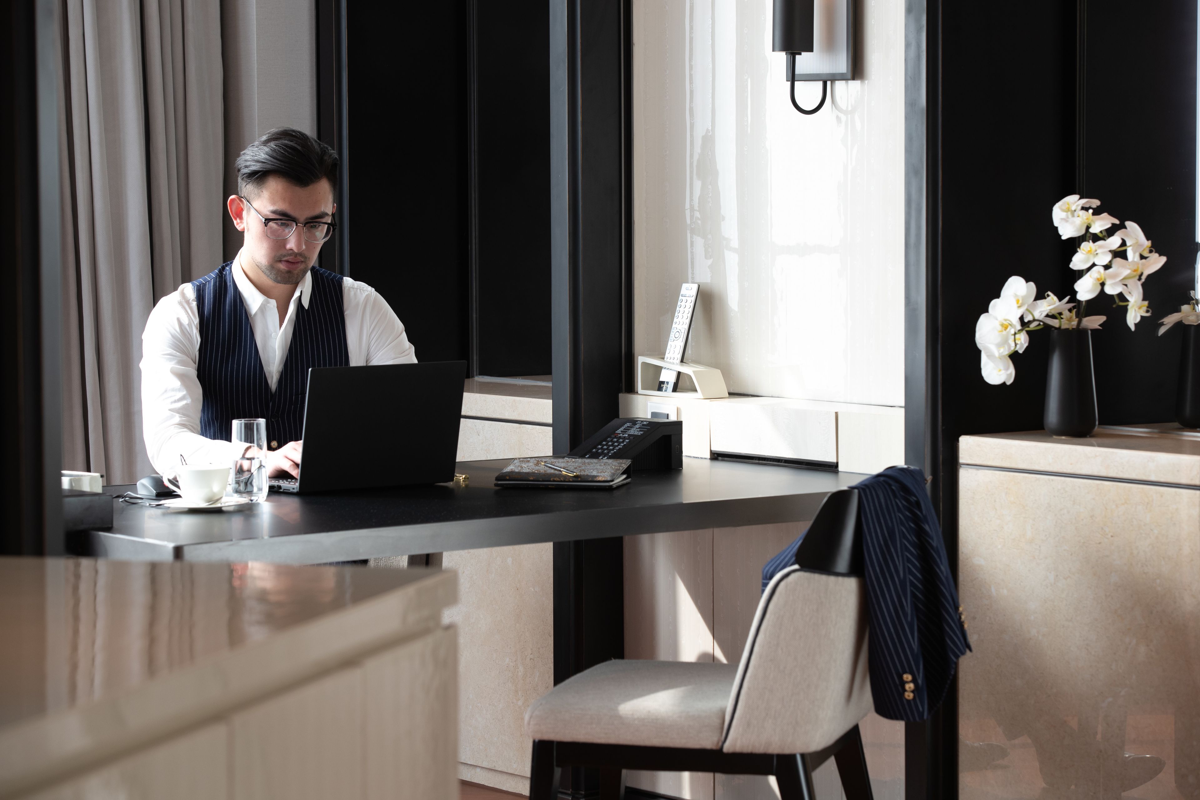 Un hombre trabajando en un escritorio con una computadora portátil, un teléfono y una taza de café. Hay un jarrón con flores blancas sobre la mesa.