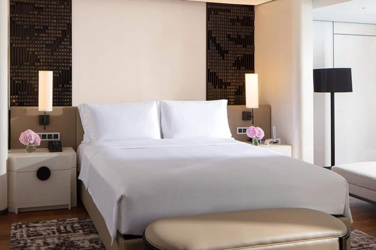Habitación de hotel con una cama grande, dos mesitas de noche con lámparas, una pequeña mesa con un florero y un banco al pie de la cama.