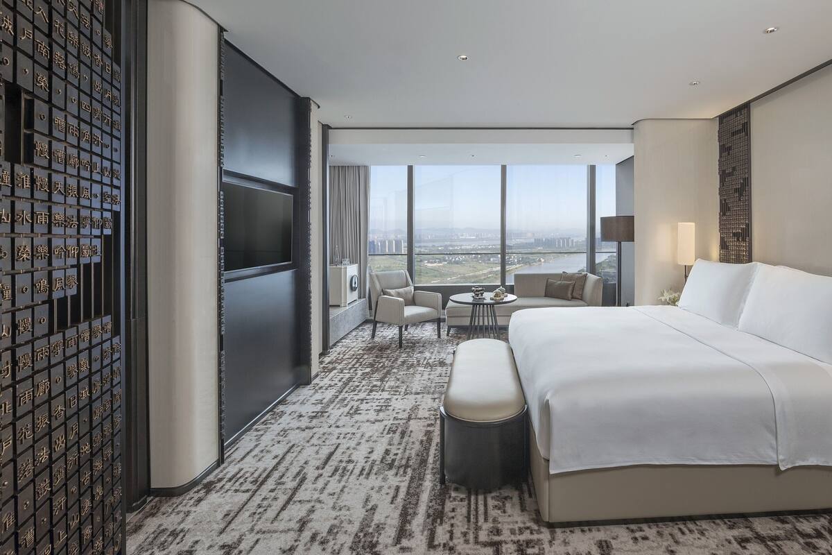 Habitación de hotel moderna con una cama grande, una zona de estar con mesa y vistas a la ciudad a través de amplias ventanas.