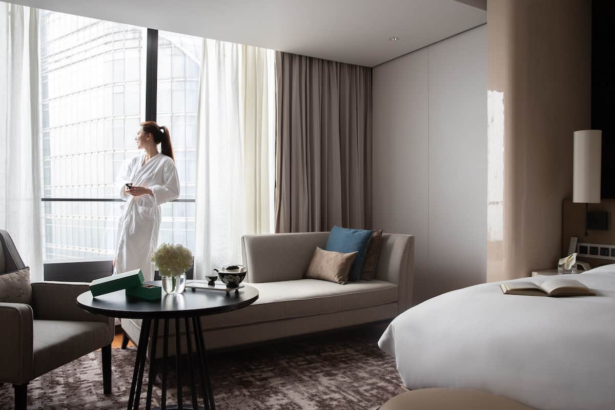 Habitación de hotel con un ventanal grande, un sofá, una mesa redonda con un florero y una tetera, y una cama con un libro abierto.