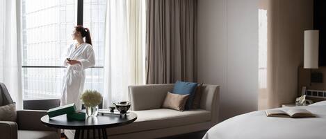 A hotel room with a large window, a sofa, a round table with a vase and a teapot, and a bed with an open book.