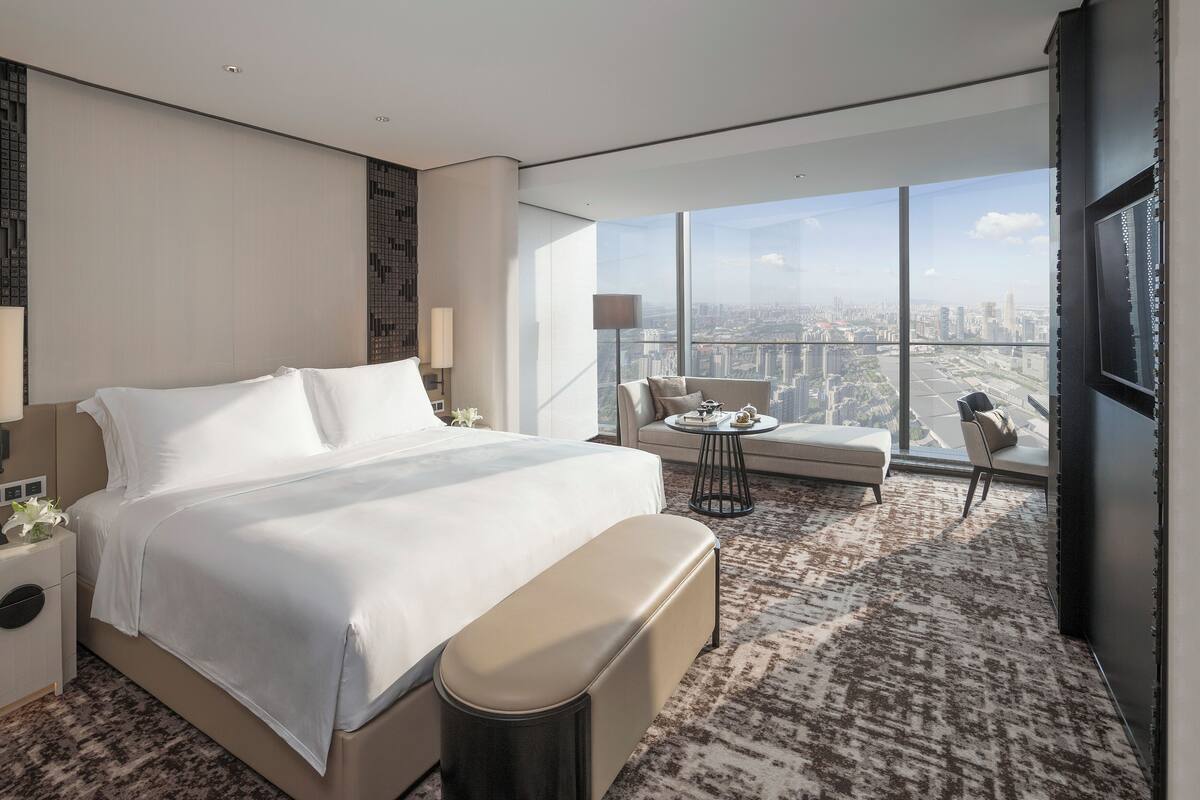 Habitación de hotel moderna con una cama grande, un sofá, una mesita y vistas a la ciudad a través de amplios ventanales.