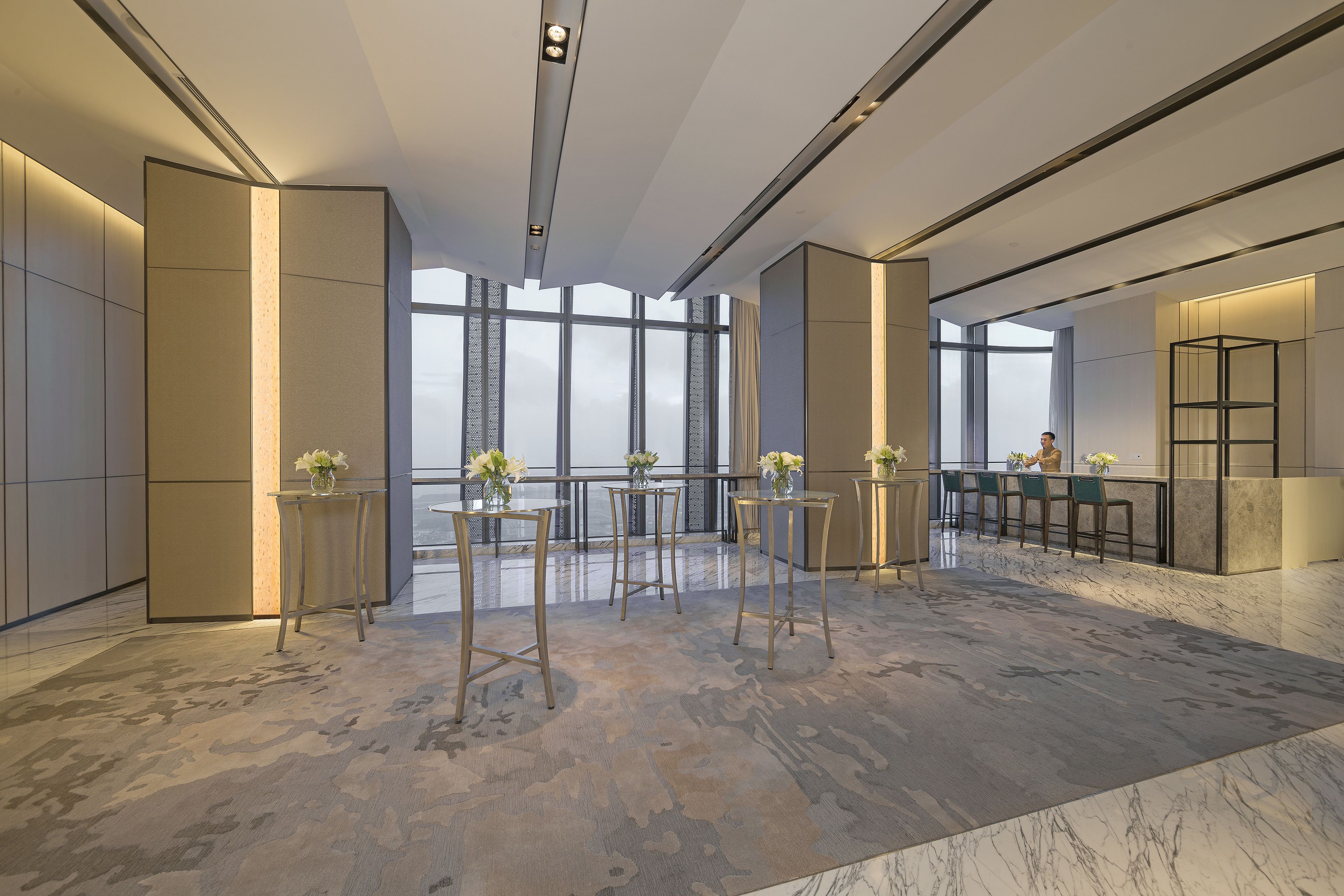 Un'elegante hall d'hotel dal design moderno, con soffitti alti, ampie finestre e un pavimento in marmo. Si notano diversi tavoli con sedie e vasi decorativi.
