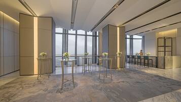 Un'elegante hall d'hotel dal design moderno, con soffitti alti, ampie finestre e un pavimento in marmo. Si notano diversi tavoli con sedie e vasi decorativi.