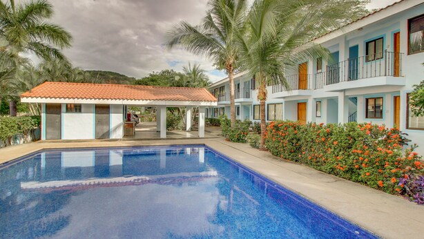 A swimming pool with a tiled deck, surrounded by a building with balconies and a covered area.
