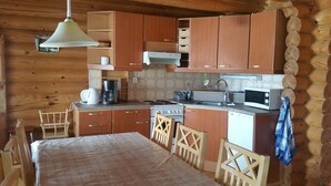 A cozy cabin-style kitchen with wooden cabinets, a stove, a sink, and a microwave. There is a dining table with chairs and a hanging light fixture. - Raikala (Savonlinna)
