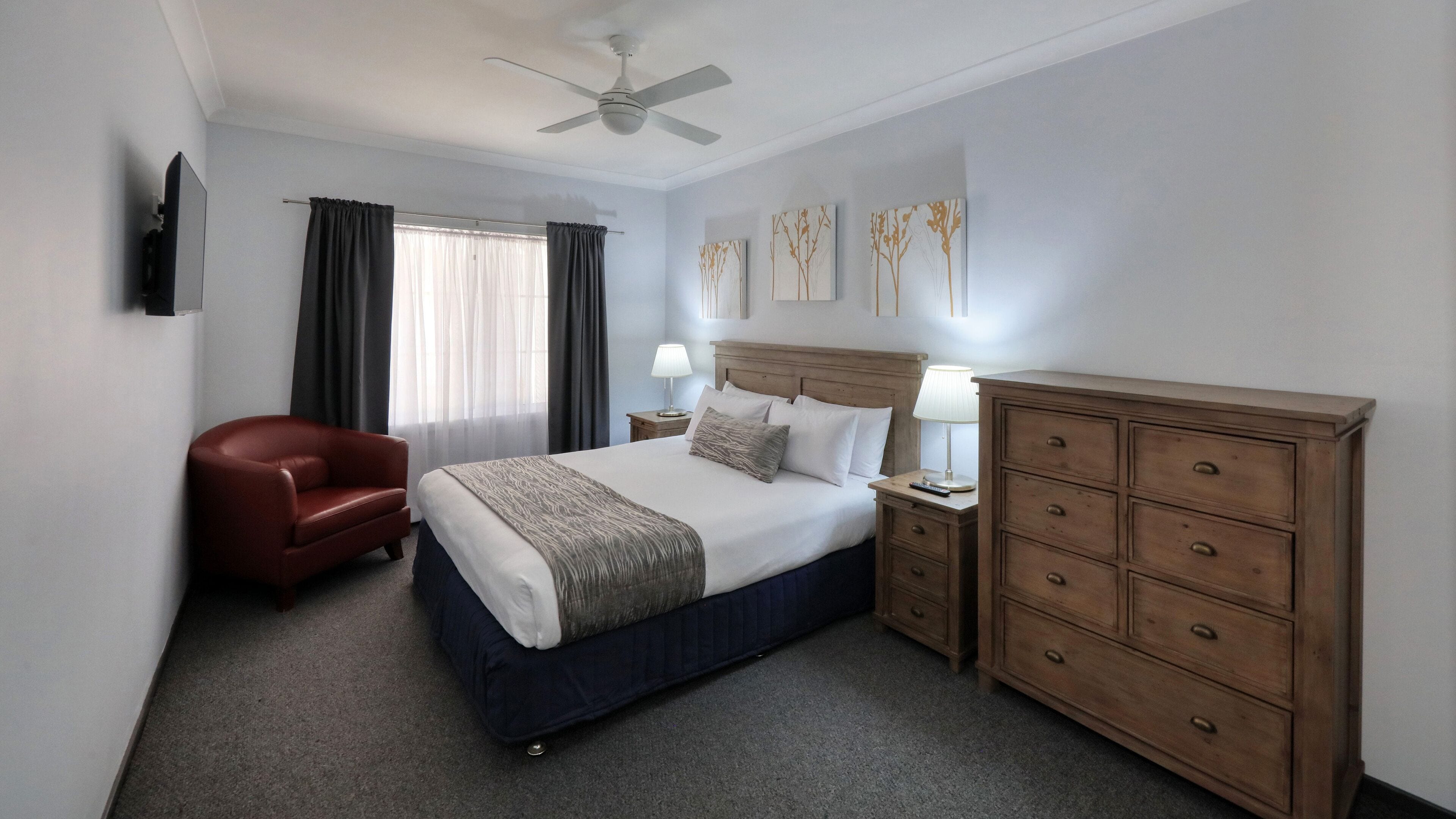 A hotel room with a bed, a wooden dresser, a red armchair, a ceiling fan, and artwork on the wall.