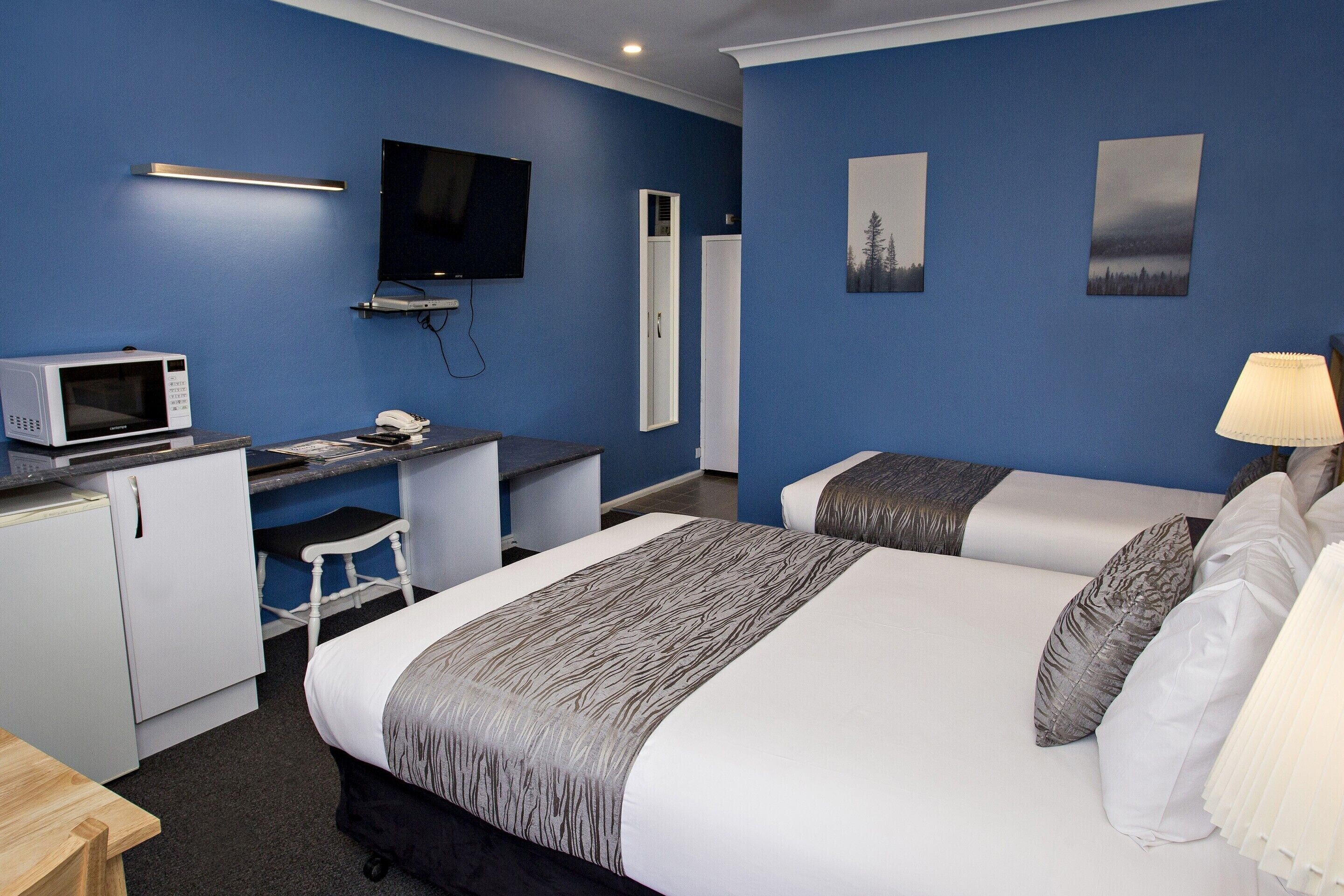 A hotel room with a bed, two bedside tables, a desk with a computer and a microwave, a TV, and artwork on the walls.
