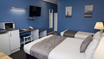A hotel room with a bed, two bedside tables, a desk with a computer and a microwave, a TV, and artwork on the walls.