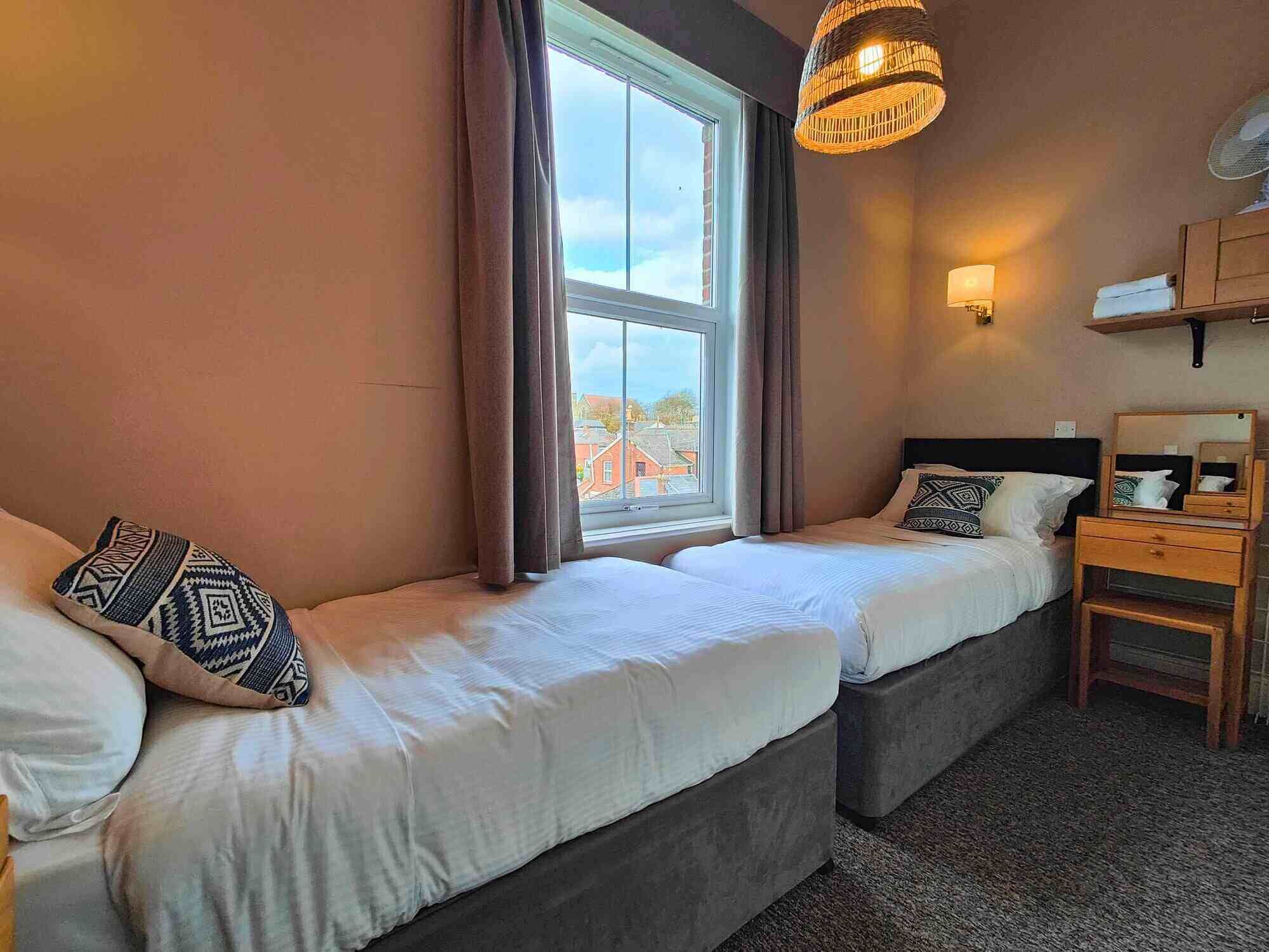 A hotel room with two beds, a window with curtains, a bedside lamp, a wooden nightstand, and a hanging light fixture.