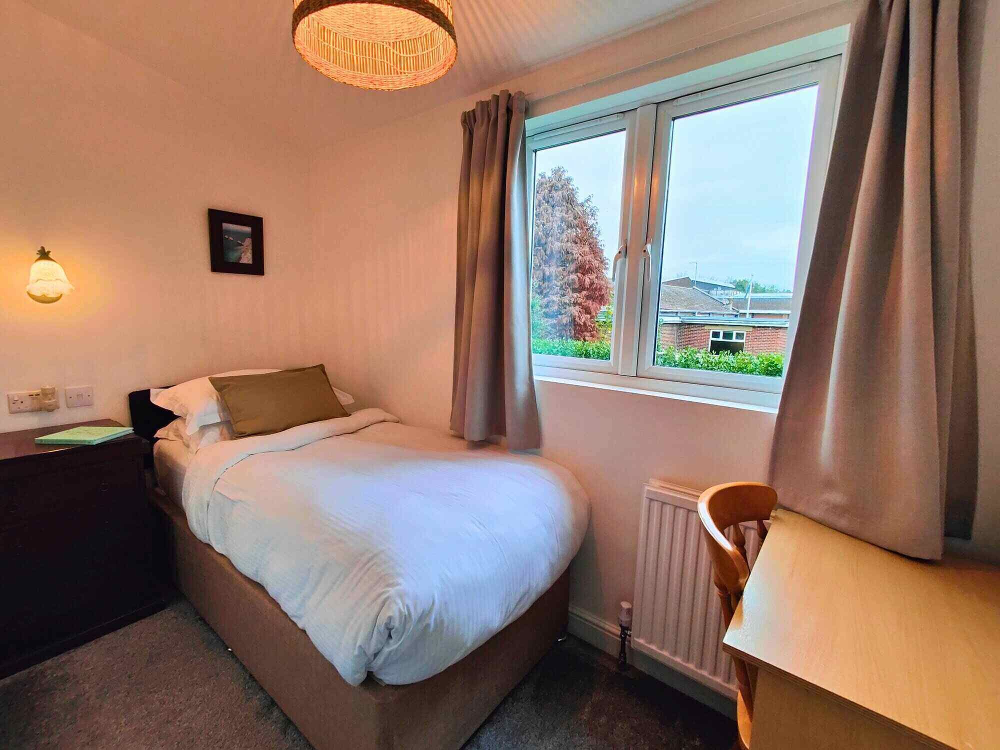 A hotel room with a bed, a desk, a chair, a window with curtains, a wall-mounted lamp, and a radiator.