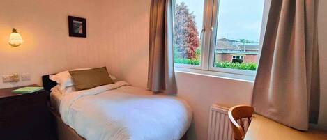 A hotel room with a bed, a desk, a chair, a window with curtains, a wall-mounted lamp, and a radiator.