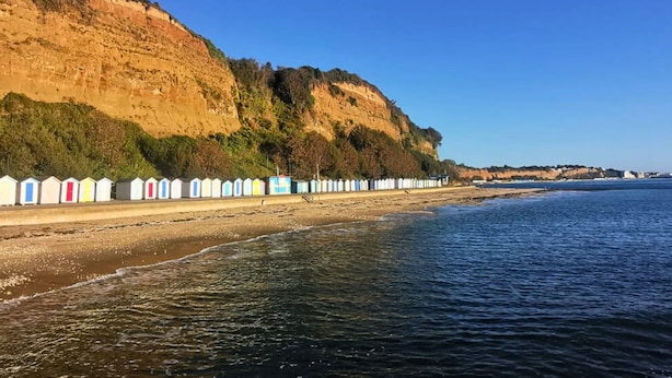 Une plage côtière bordée d’une rangée de cabanes colorées, une falaise couverte de végétation et un ciel bleu dégagé.