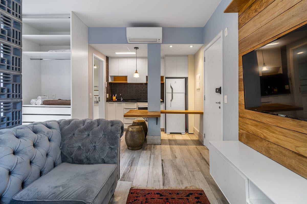 Un salon moderne avec un canapé gris, un mur d’accent en bois et un coin cuisine à l’arrière-plan.
