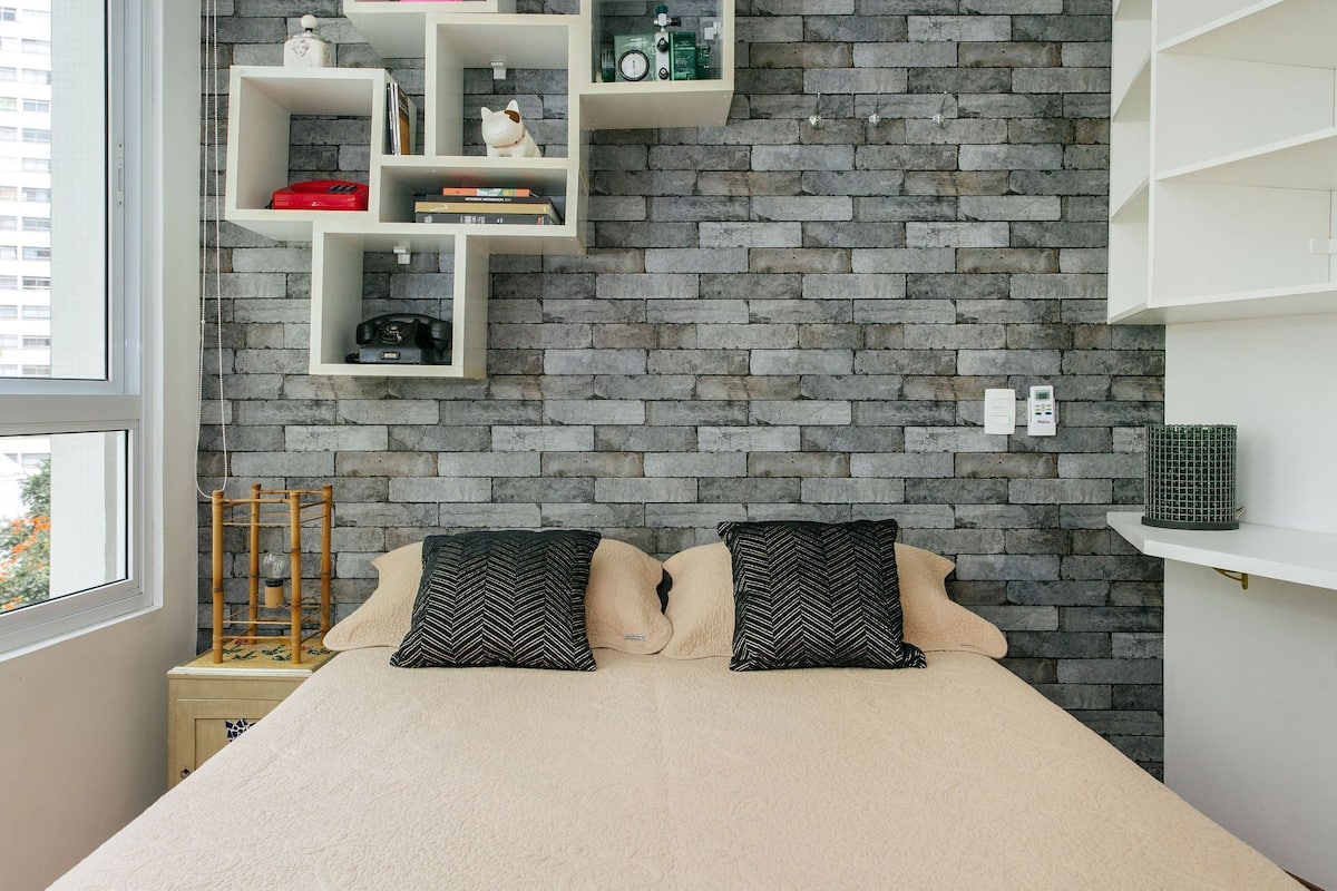 Une chambre avec un mur en briques, un lit avec deux oreillers, une table de chevet et une fenêtre.