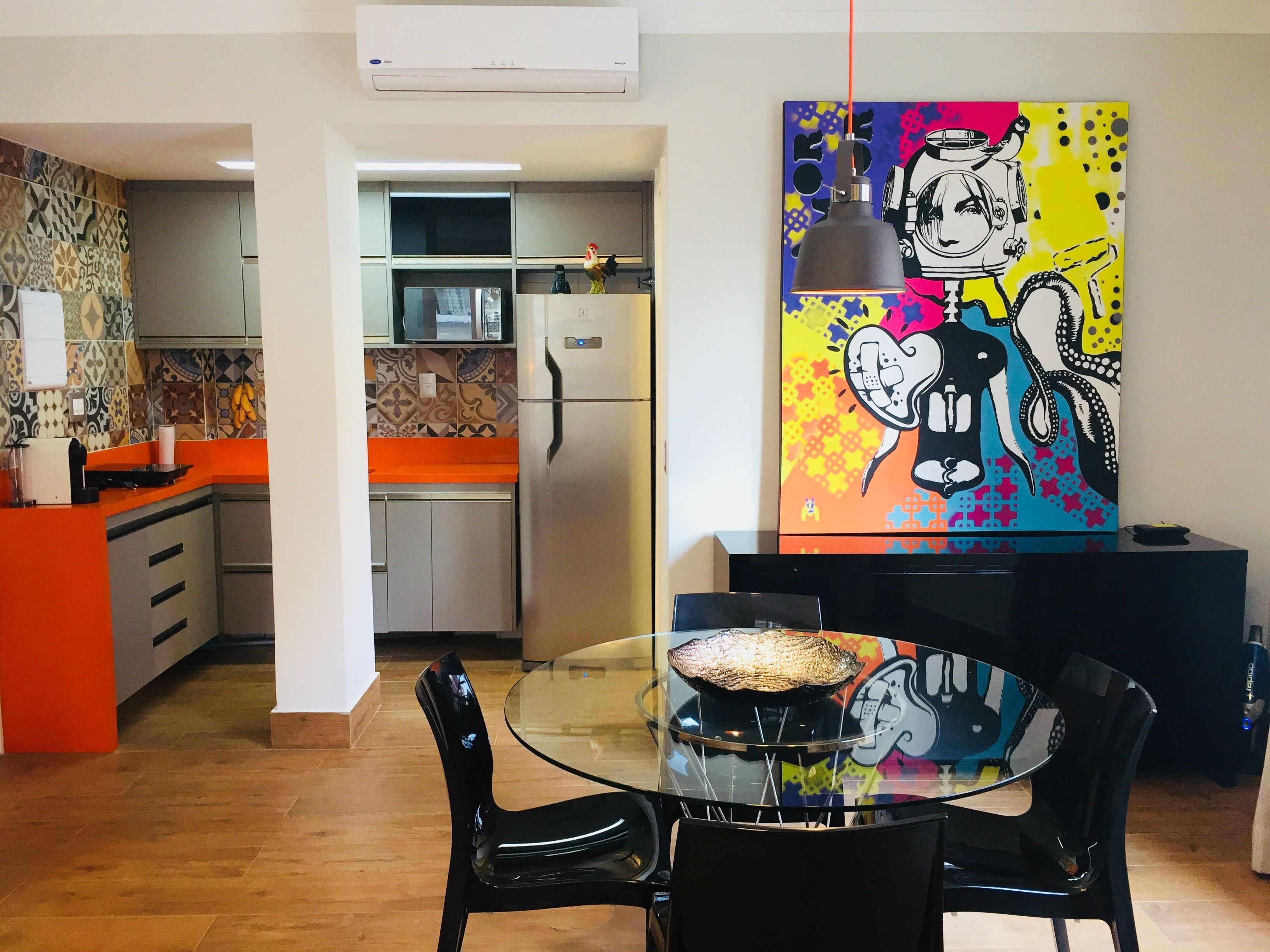 A modern kitchen with a refrigerator, microwave, and orange countertop. There is a dining area with a glass table and black chairs. A colorful painting is on the wall.