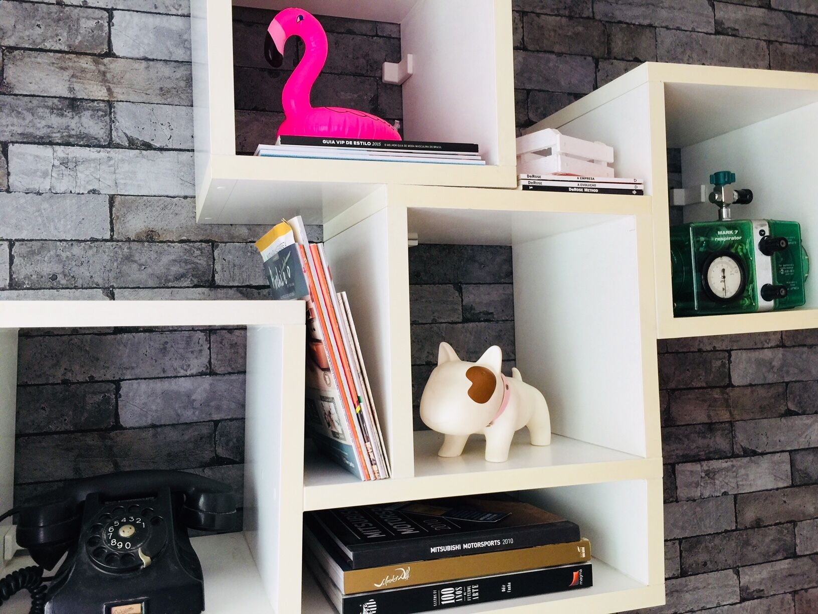 A bookshelf with various items including books, a pink flamingo, a piggy bank, a green respirator, and a black rotary phone.