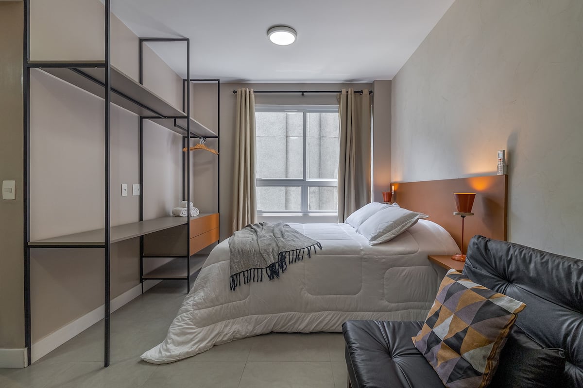 Une chambre à coucher avec un lit, un canapé en cuir noir, une étagère fixée au mur et une fenêtre avec des rideaux.