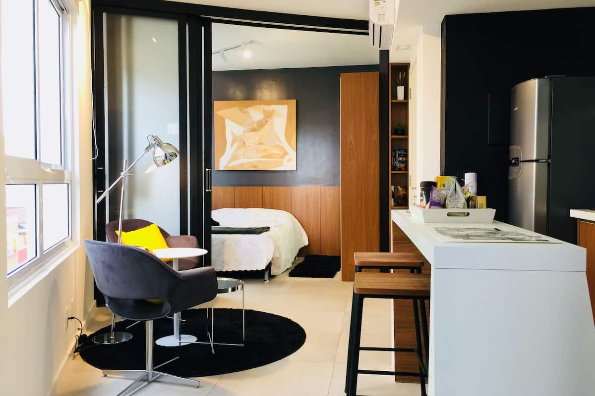Une chambre d’hôtel moderne équipée d’un lit, d’un bureau, d’une chaise, d’un petit coin cuisine et d’une grande fenêtre.