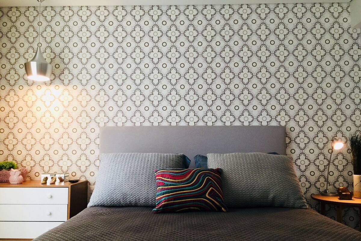 Une chambre à coucher bien aménagée, avec un papier peint à motifs, une tête de lit grise et un coussin rayé aux couleurs vives.