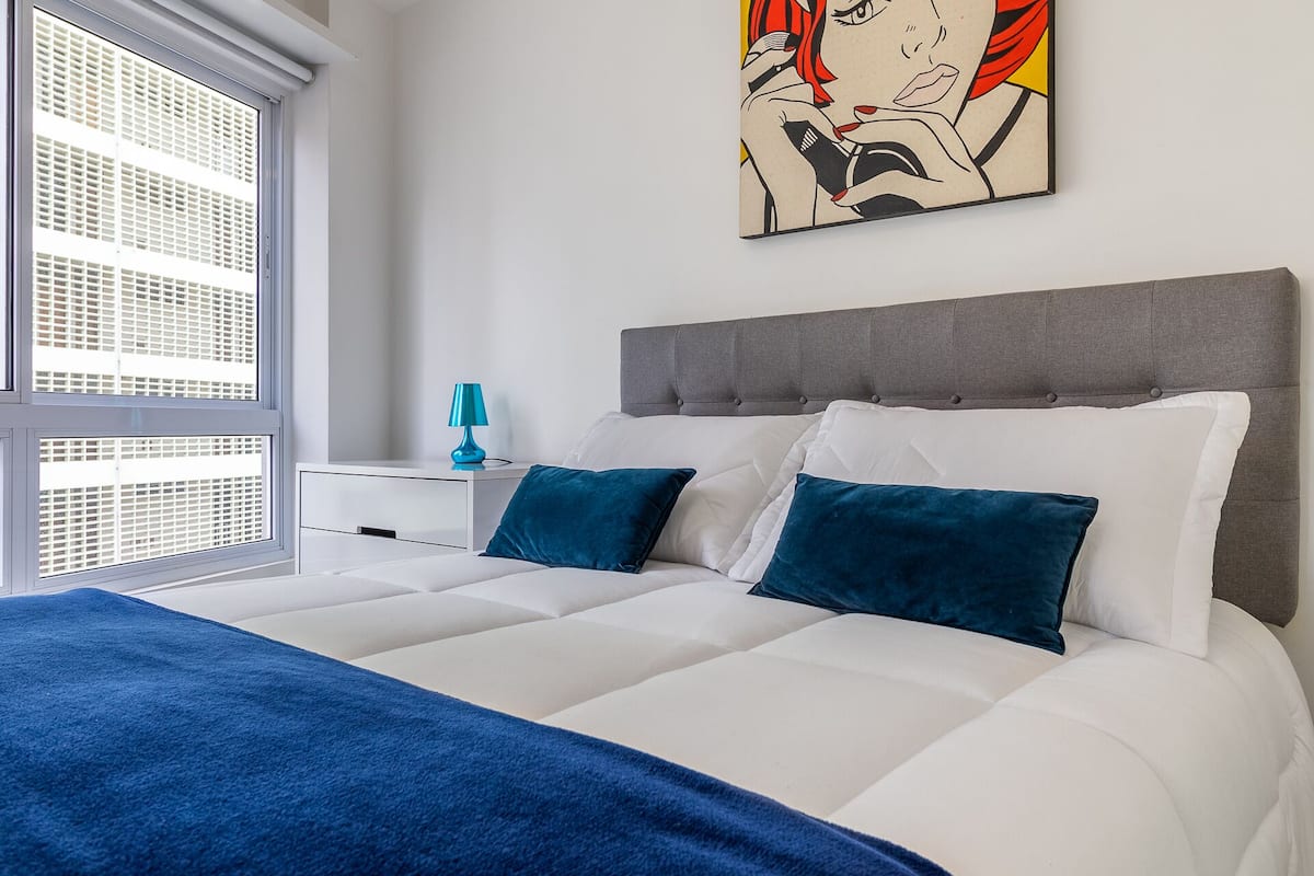 Une chambre moderne avec un grand lit, une tête de lit rembourrée de couleur grise et une fenêtre avec des stores.
