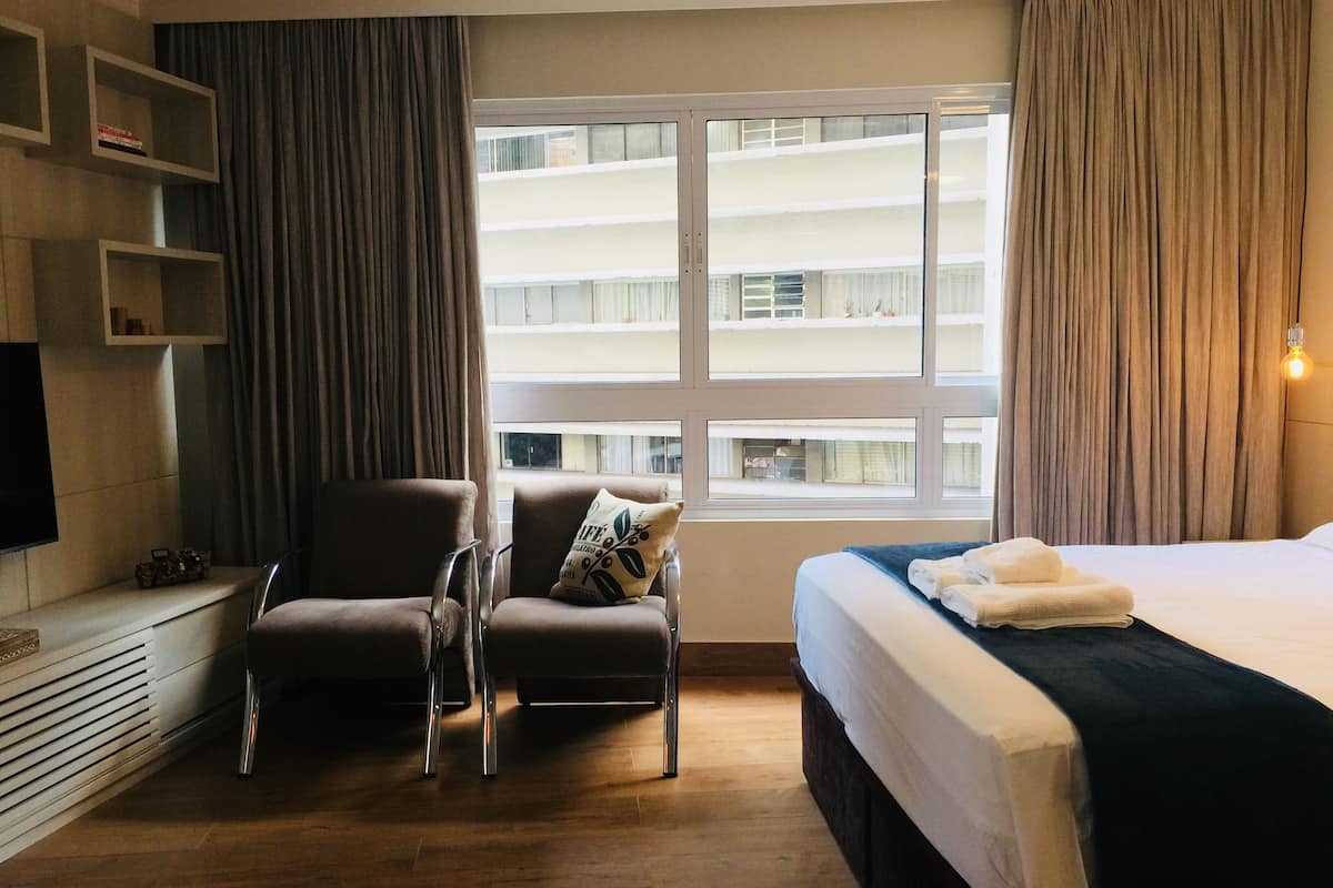 Une chambre d’hôtel avec un lit, une chaise, une télévision et une fenêtre donnant sur des immeubles.