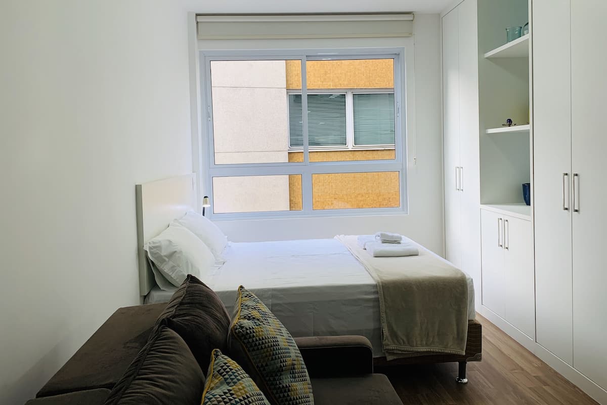 Une chambre à coucher comprenant un lit, une table de chevet, une lampe, une fenêtre avec des stores, une armoire et un canapé.