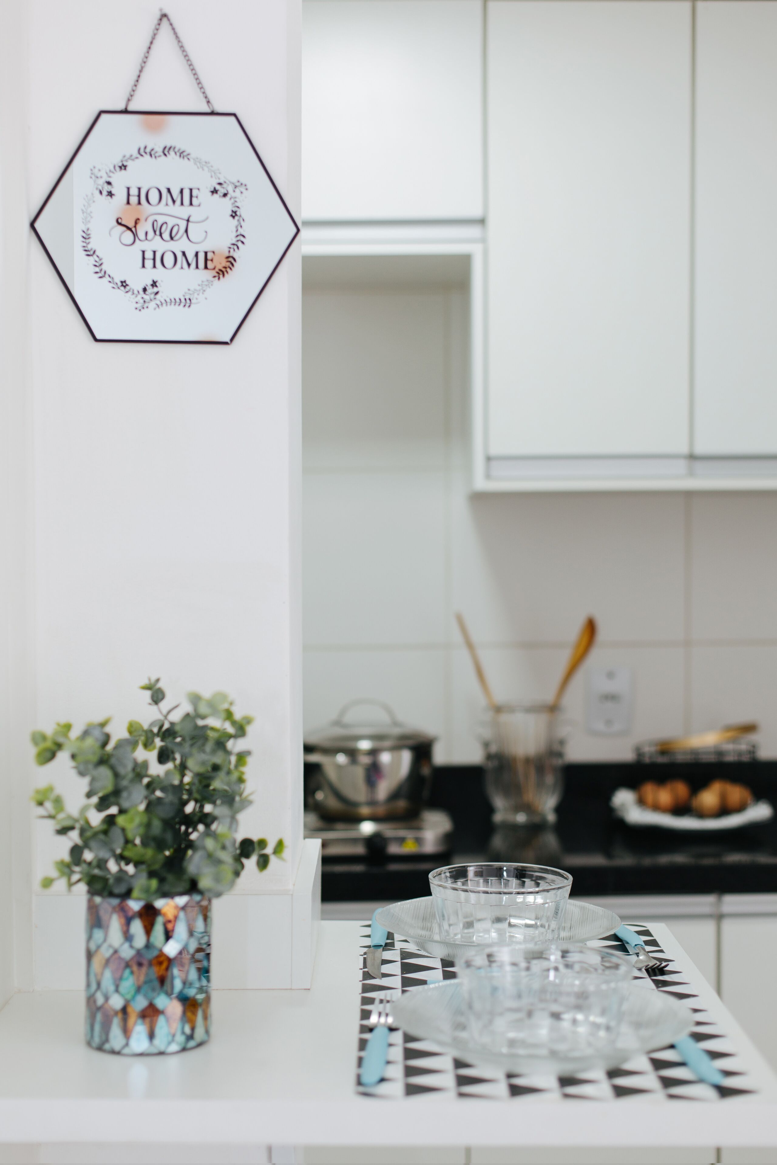 A kitchen with white cabinets, a hexagonal sign on the wall, a potted plant, and a table with glasses and cutlery.