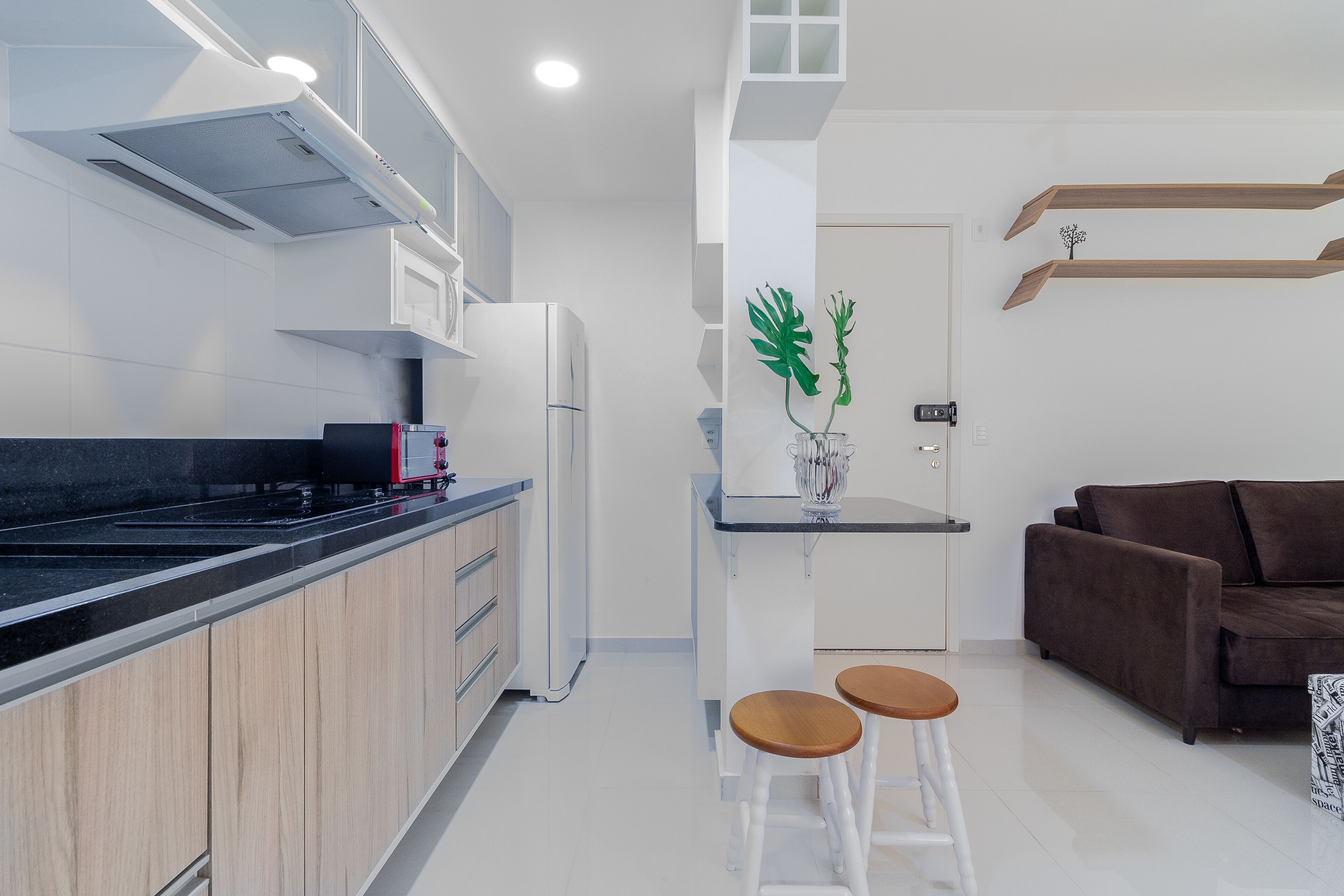 Eine moderne Küche mit weißen Schränken, einer schwarzen Arbeitsplatte und einem eingebauten Herd. Im Wohnbereich befinden sich zwei Hocker und ein braunes Sofa.
