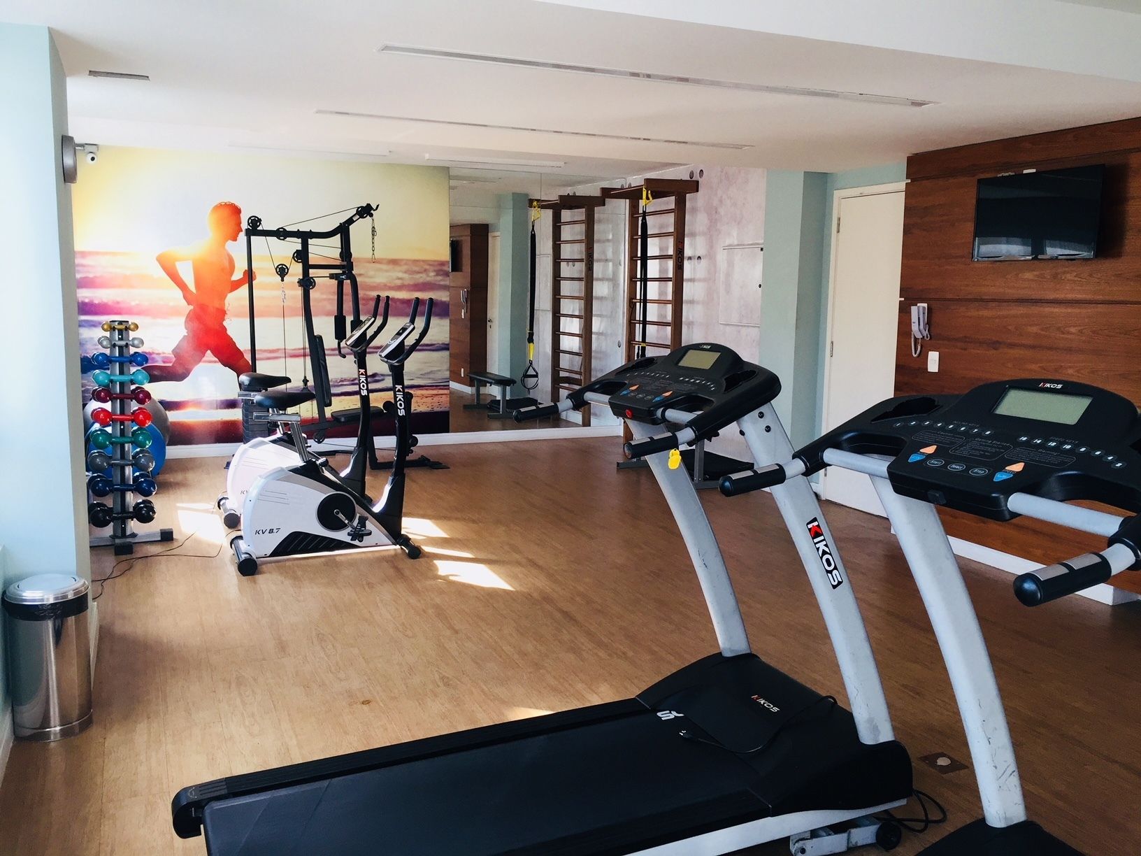 Een sportschool met cardioapparatuur, waaronder een loopband en hometrainers, en een spiegelwand.