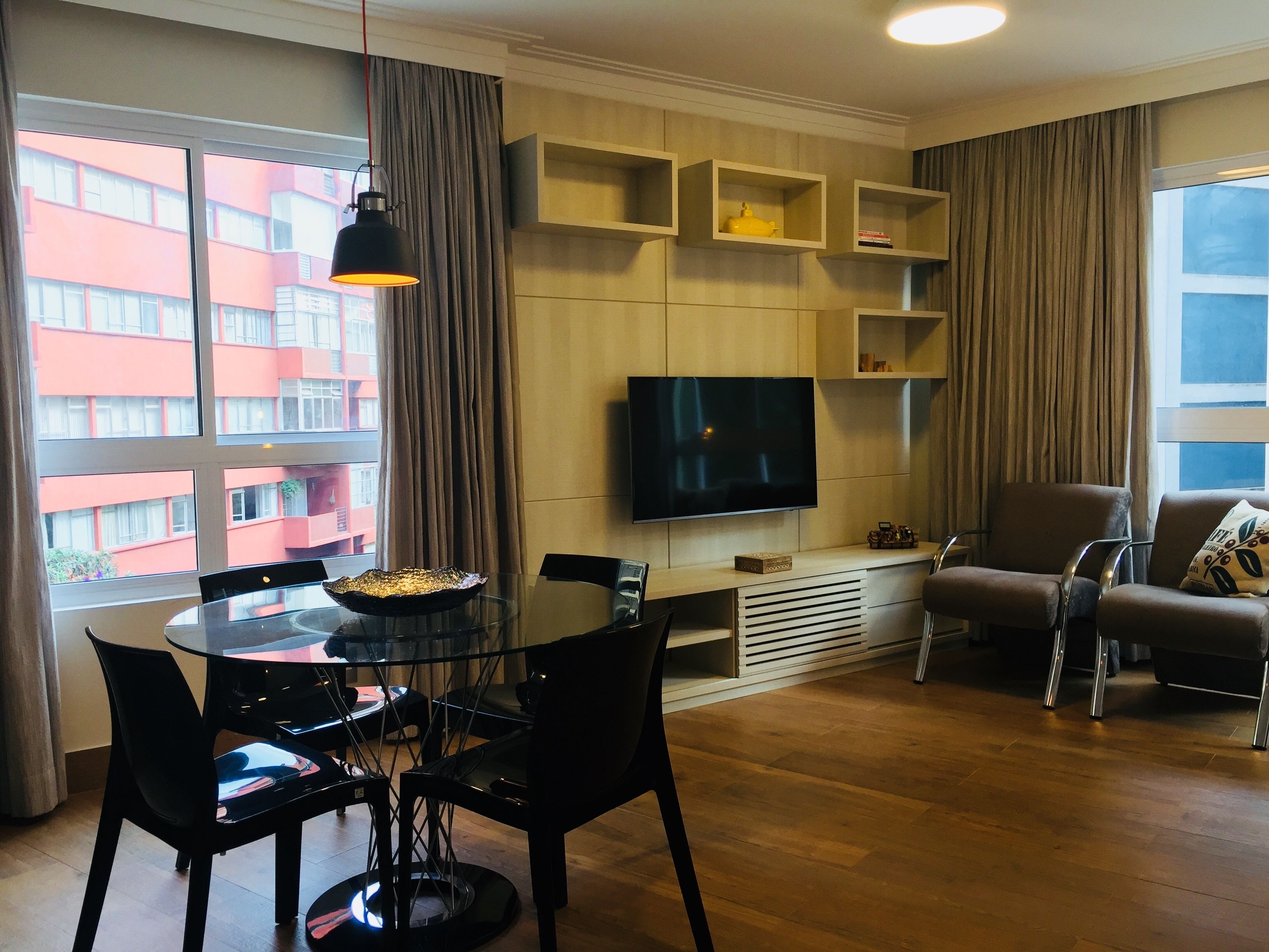 Uma sala de estar moderna com televisão de tela plana, uma mesa de jantar com cadeiras, um sofá e uma janela grande com cortinas.