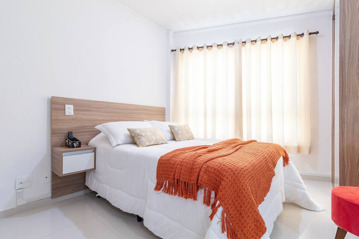 Une chambre moderne avec une tête de lit en bois, une literie blanche, un plaid rouge et une table de chevet avec une télécommande.