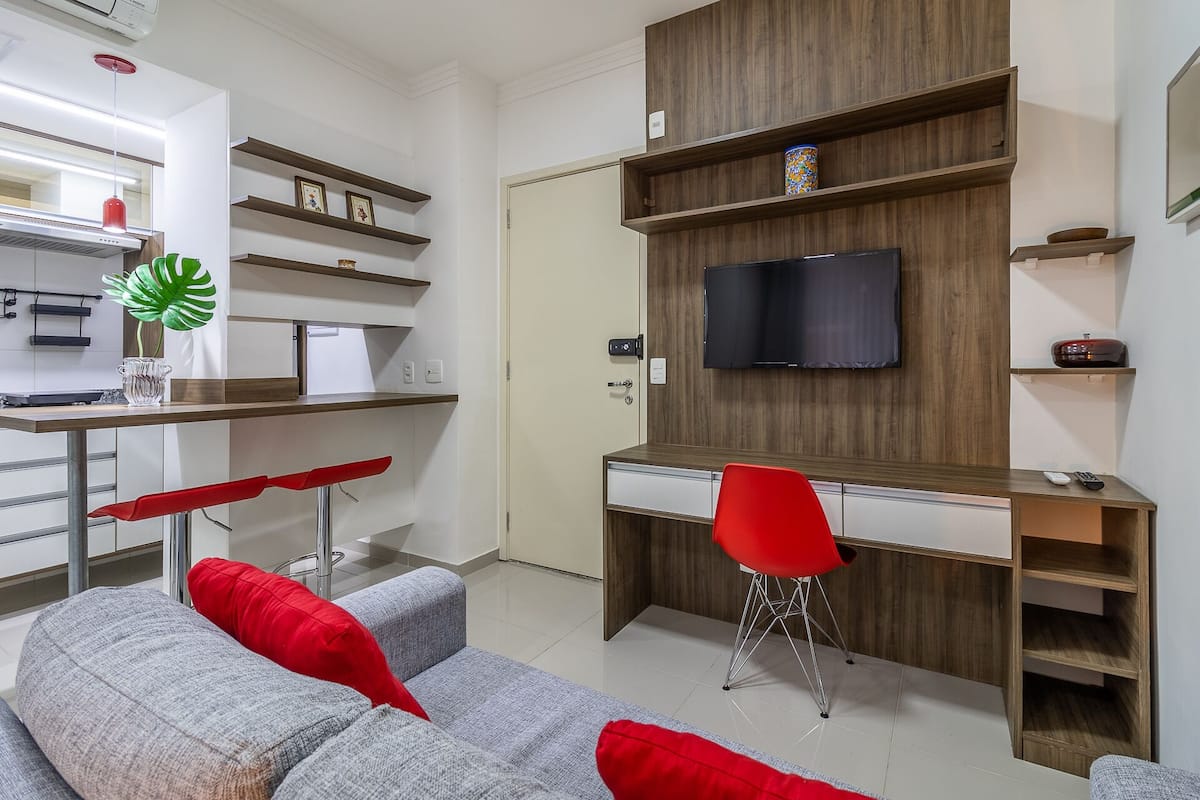 Un salon moderne avec un canapé gris, des coussins rouges, un bureau en bois, une chaise rouge et un téléviseur fixé au mur.