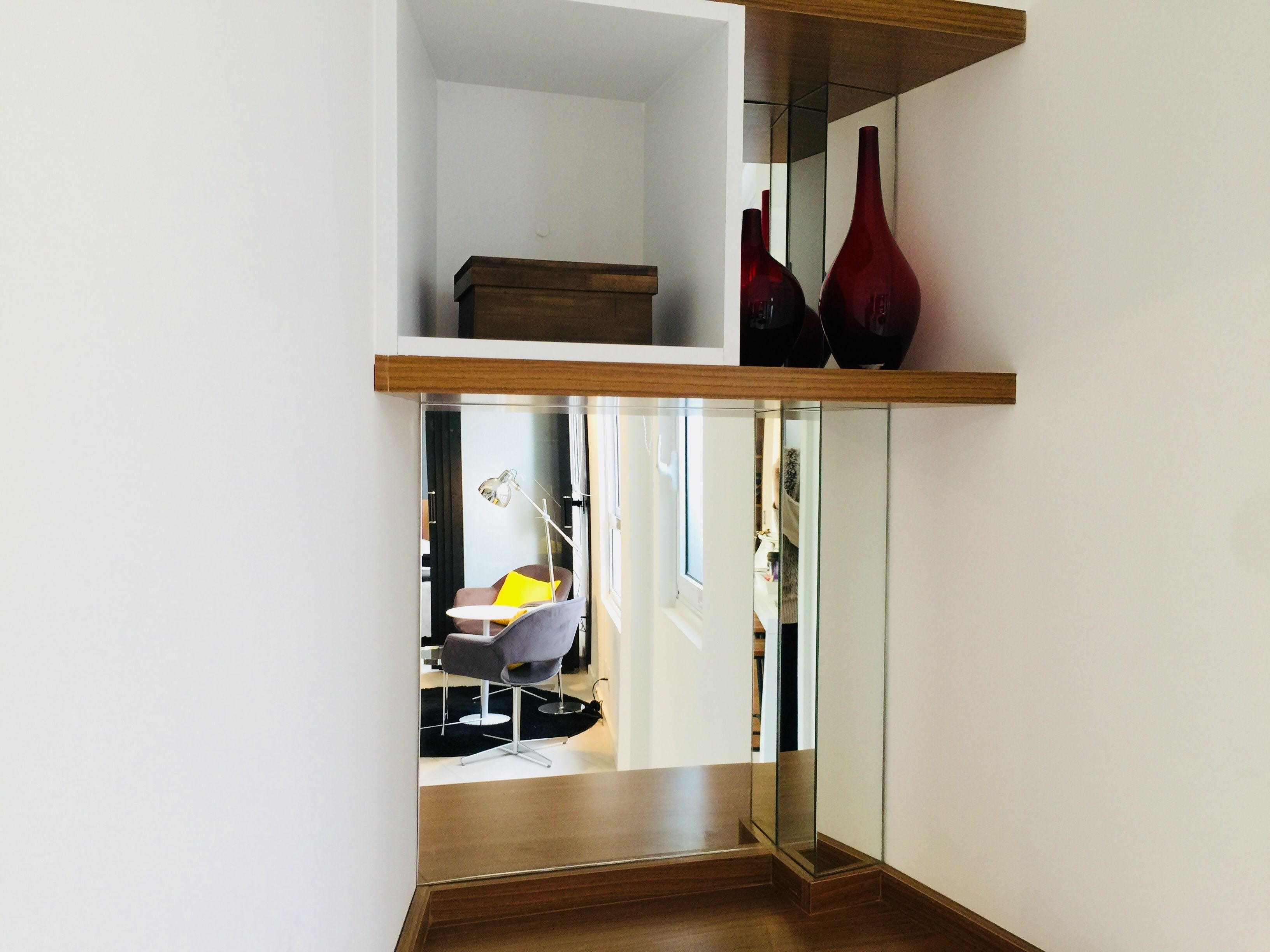 A modern interior with a wooden floor, a glass door leading to a room with a chair and a lamp, and a shelf with two red vases.