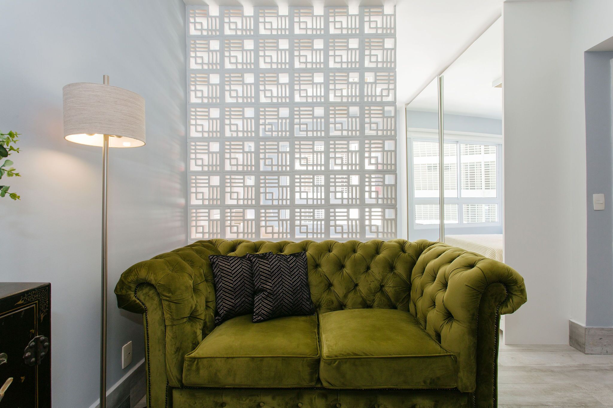 A green velvet sofa with two patterned pillows, a floor lamp, and a decorative screen with geometric patterns.