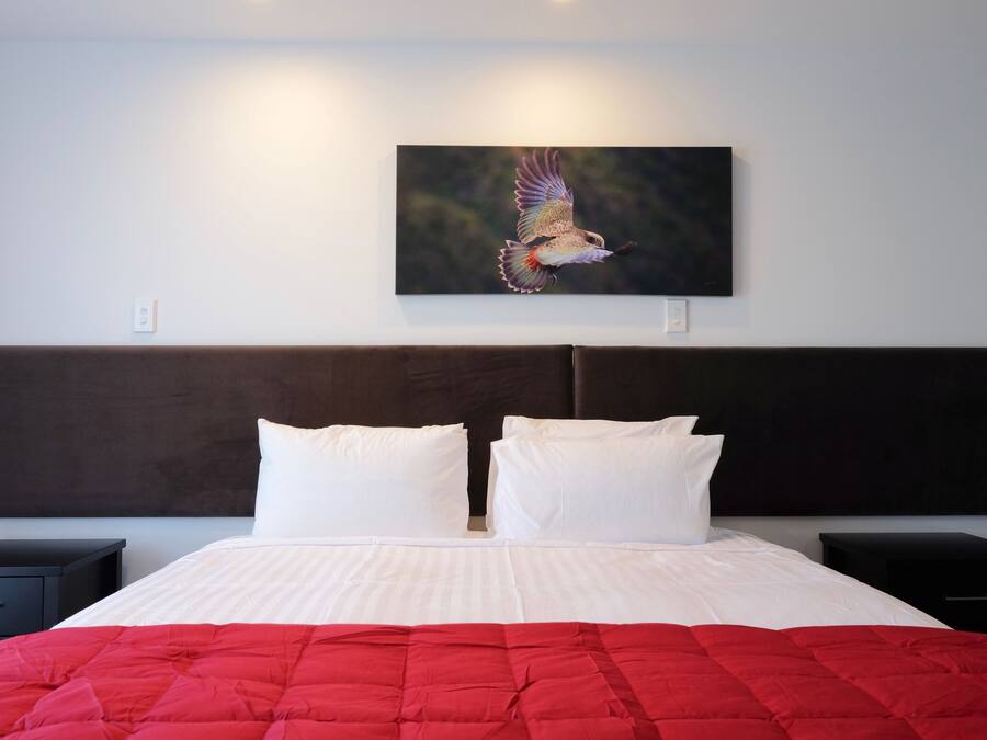 Ein ordentlich bezogenes Bett mit roter Decke, weißen Kissen und einem dunklen Kopfteil. Über dem Bett hängt ein Gemälde, das einen fliegenden Vogel zeigt.