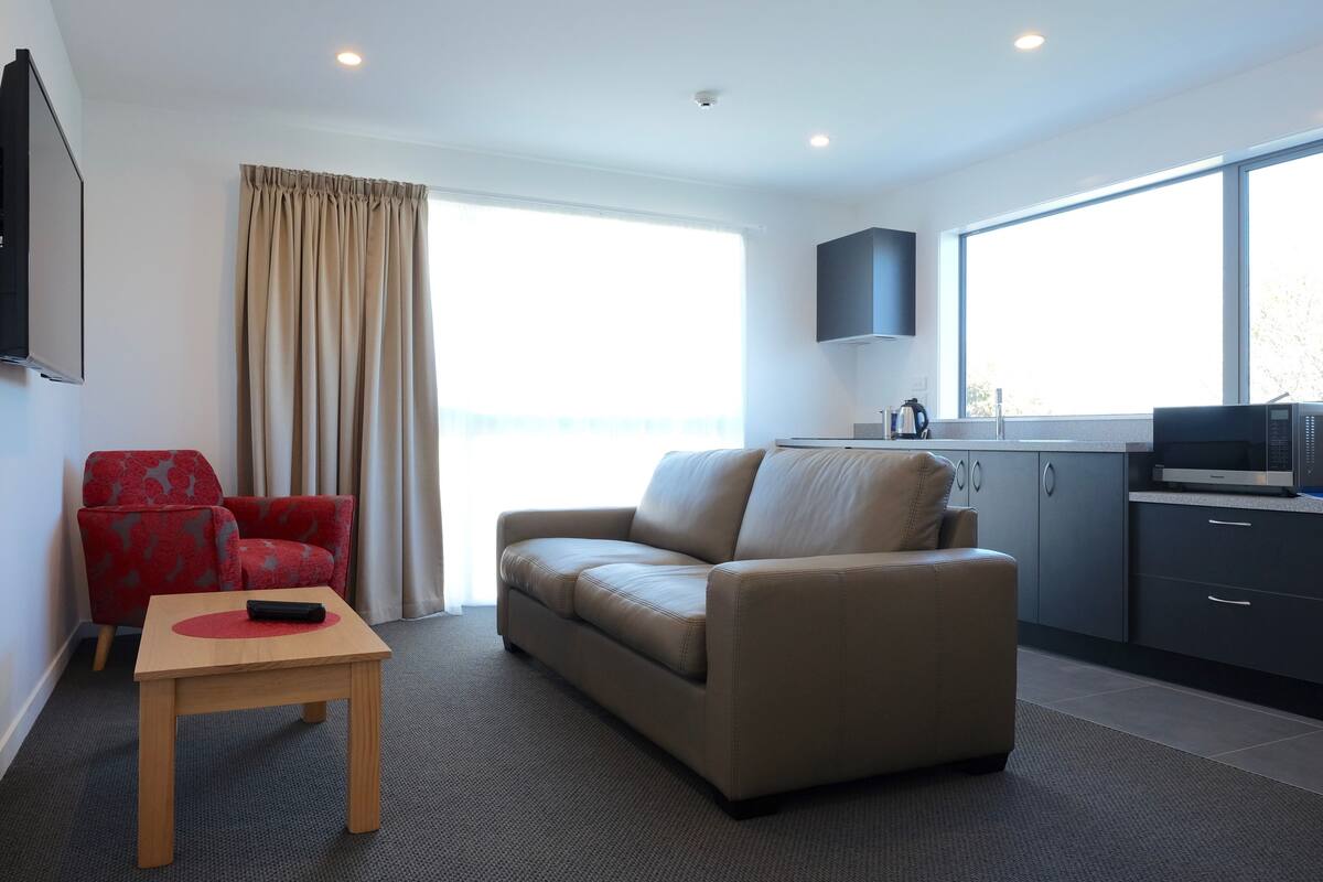 Quarto de hotel moderno com sofá bege, uma poltrona vermelha, uma pequena mesa de madeira com um controle remoto, uma área de cozinha com micro-ondas e uma janela grande que oferece luz natural.