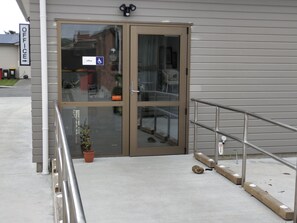 Entrada de um edifício com porta de vidro, sinal de acessibilidade para cadeiras de rodas e um pequeno vaso de planta no chão.