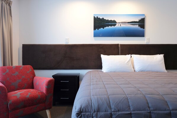 Quarto de hotel com uma cama, uma cadeira, uma mesinha de cabeceira e um quadro de um lago.