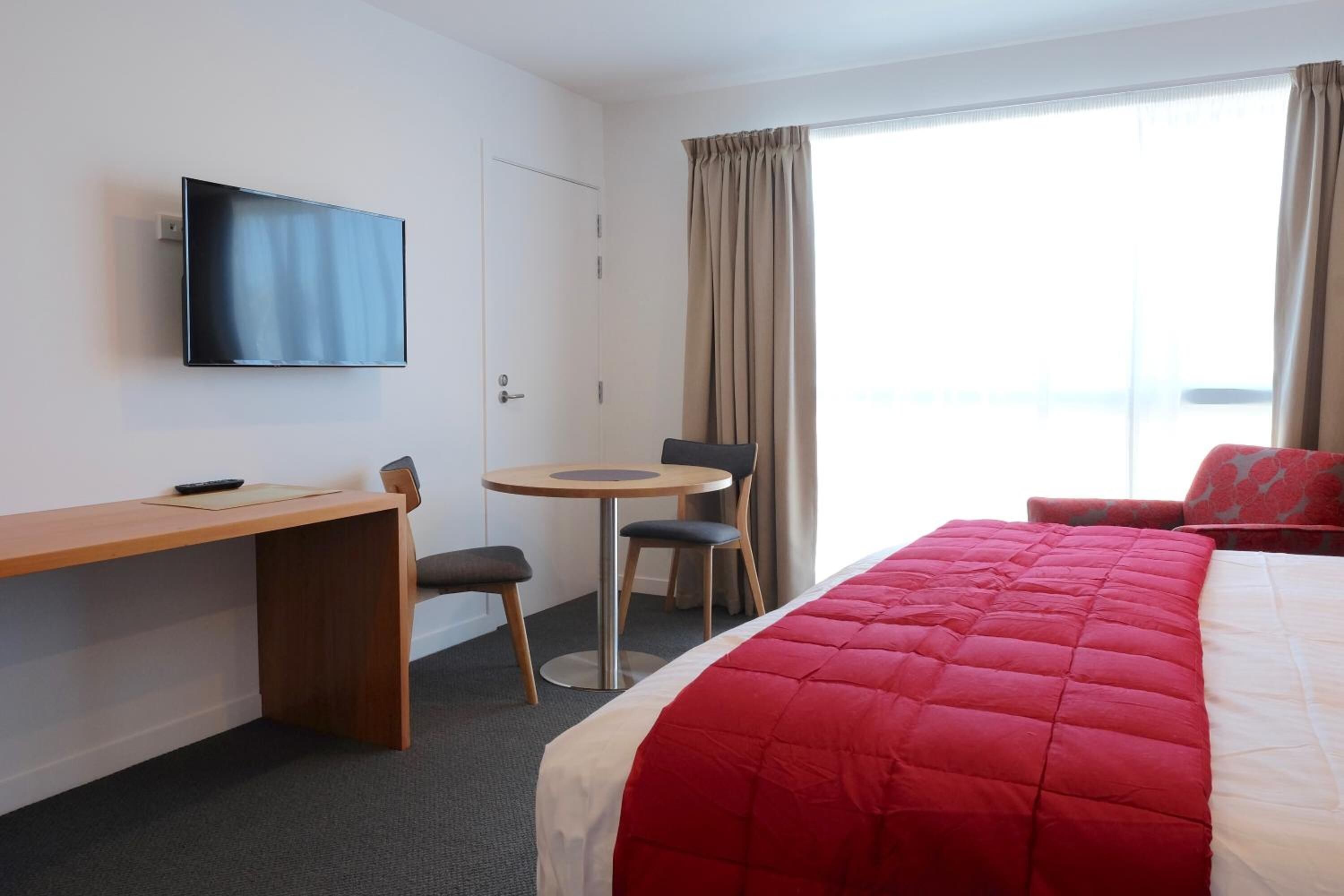 Habitación de hotel con cama, escritorio, silla, mesita, televisor montado en la pared y ventana con cortinas.