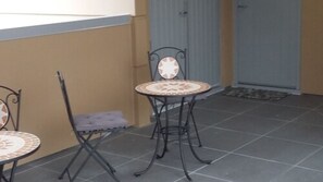 Uma pequena área externa com assentos, duas cadeiras de metal e uma mesa, uma porta ao fundo e piso de azulejos.