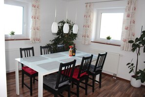 A dining area with a white table, black chairs, red cushions, hanging pendant lights, and a potted plant.