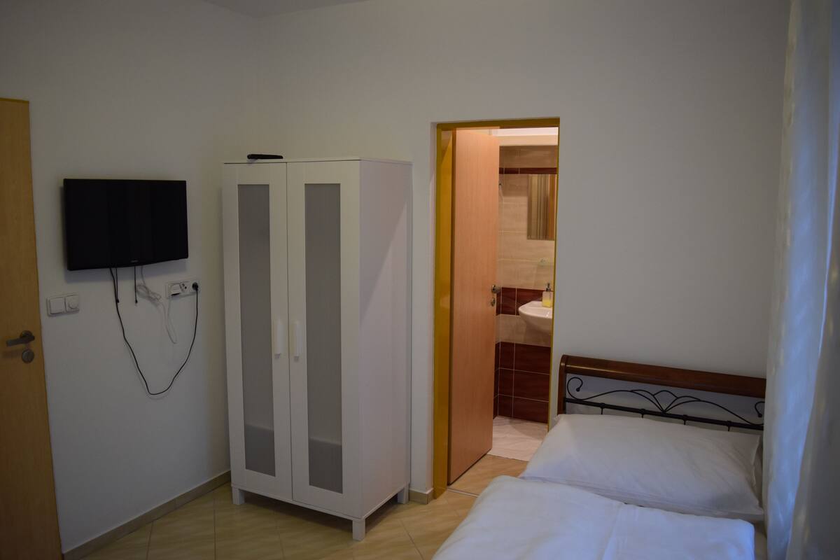 A bedroom with a bed, a wardrobe, a television mounted on the wall, and a bathroom visible through an open door.